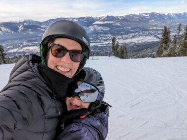 A woman takes a selfie with her young son in a snowy valley near a mountain range. Both are wearing helmets and ski jackets, and the woman is wearing sunglasses while the boy is wearing ski goggles. They're hugging each other and have big smiles. 