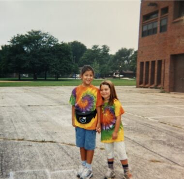Two girls in tie-dye T-shirts stand in a large, open asphalt space. The one on the left is much taller. Green grass and trees are in the distance behind them; a building is at the right.